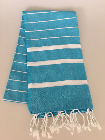 Turquoise Turkish towel peshtemal with white stripes and fringed edges, folded neatly on a light background.