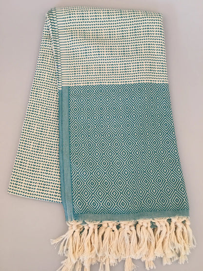 "Turquoise and cream Turkish towel with diamond pattern and fringed edges, folded on a light background."