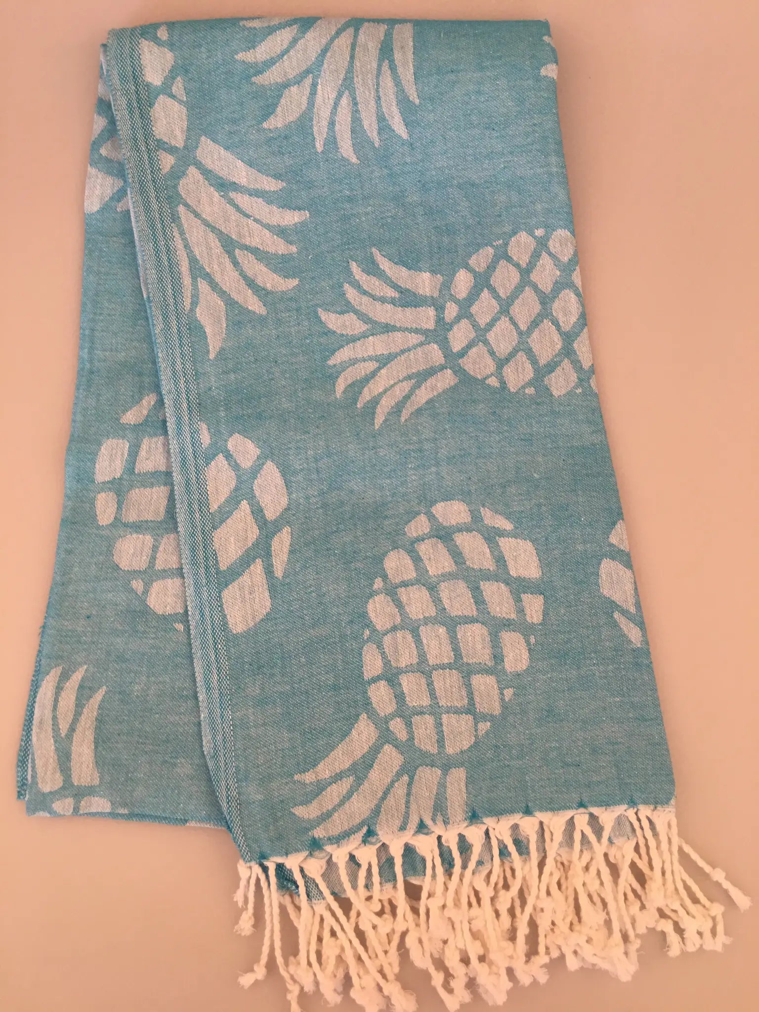 Turquoise Turkish towel with white pineapple jacquard pattern and fringed edges, laid flat on a light background.