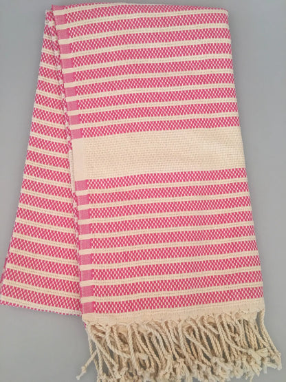 "Pink and cream striped Turkish towel with fringed edges, folded on a gray background."