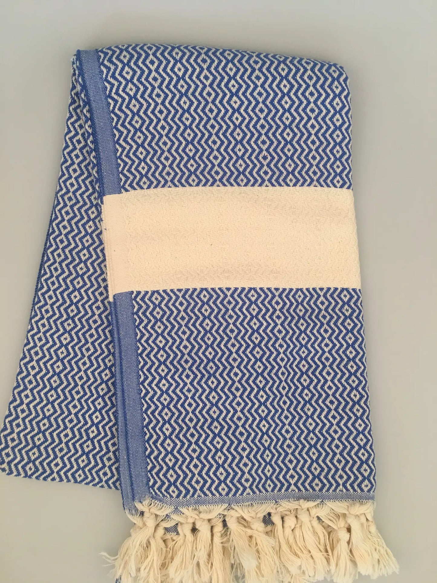 "Blue and white patterned Turkish towel with fringed edges and a solid cream band, neatly folded on a light surface."