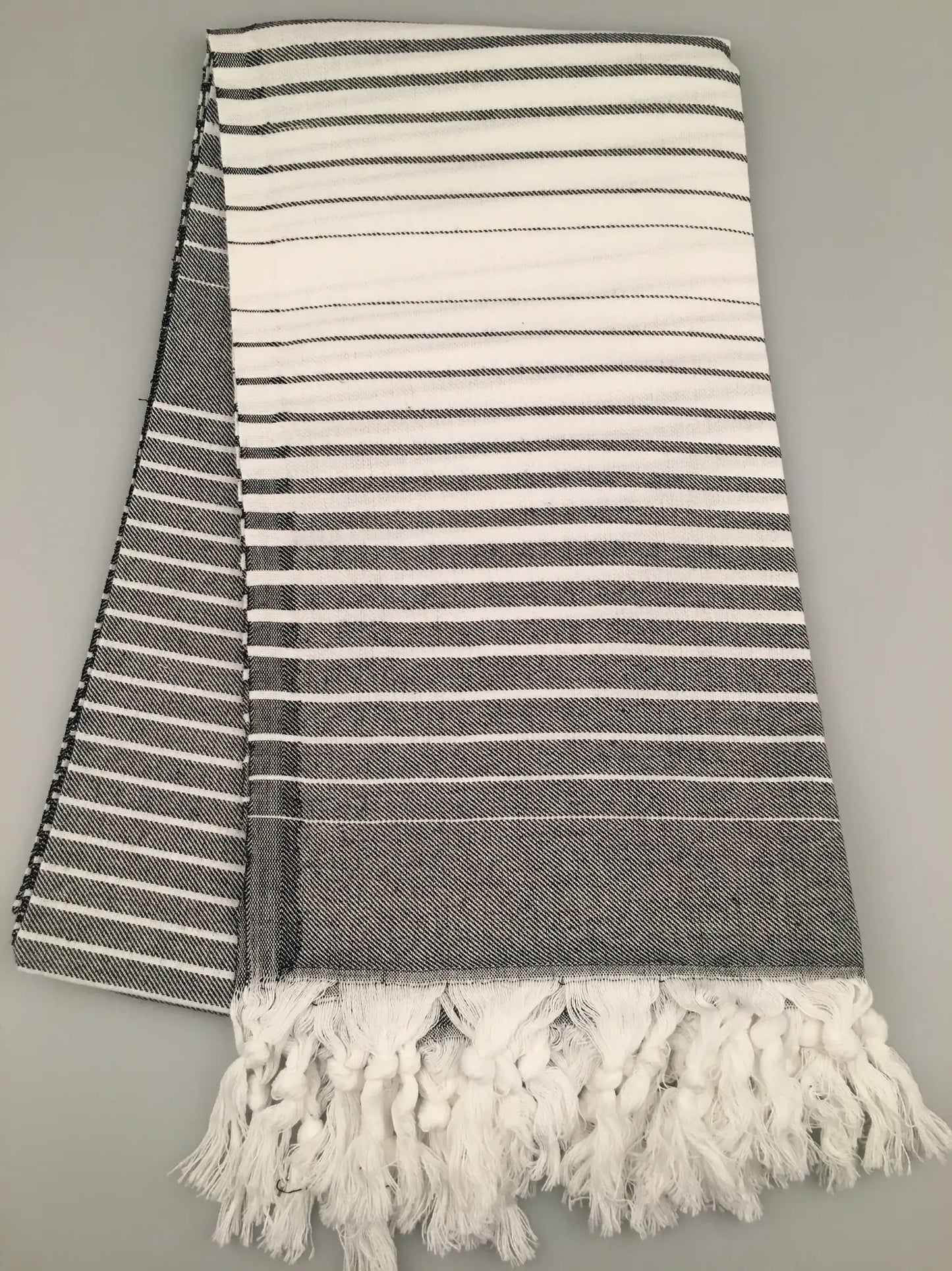 "Telmessos Turkish towel peshtemal in gray and white stripes, featuring fringed edges, laid flat on a gray background."