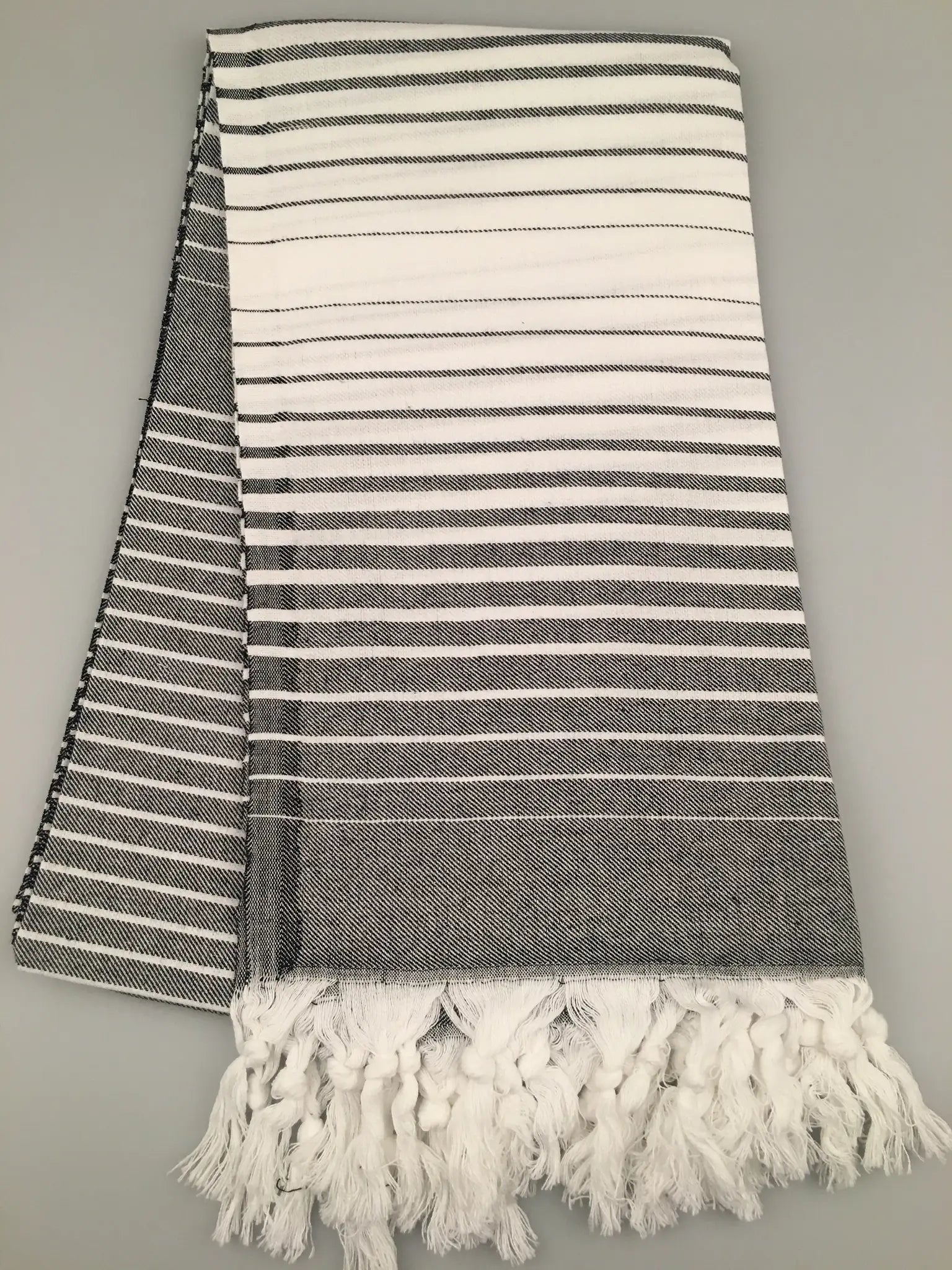 "Telmessos Turkish towel peshtemal in gray and white stripes, featuring fringed edges, laid flat on a light background."