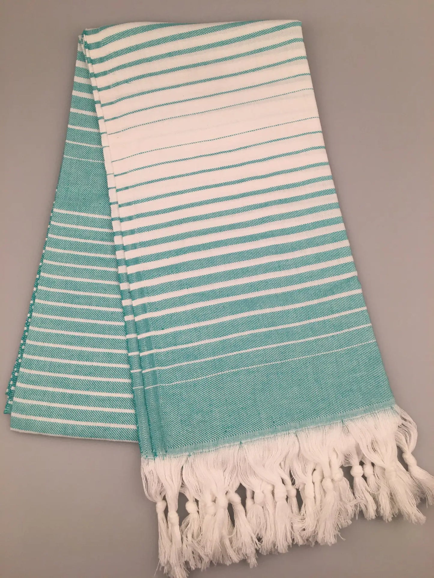 Turquoise and white striped Turkish towel with fringed edges, laid flat on a gray background.