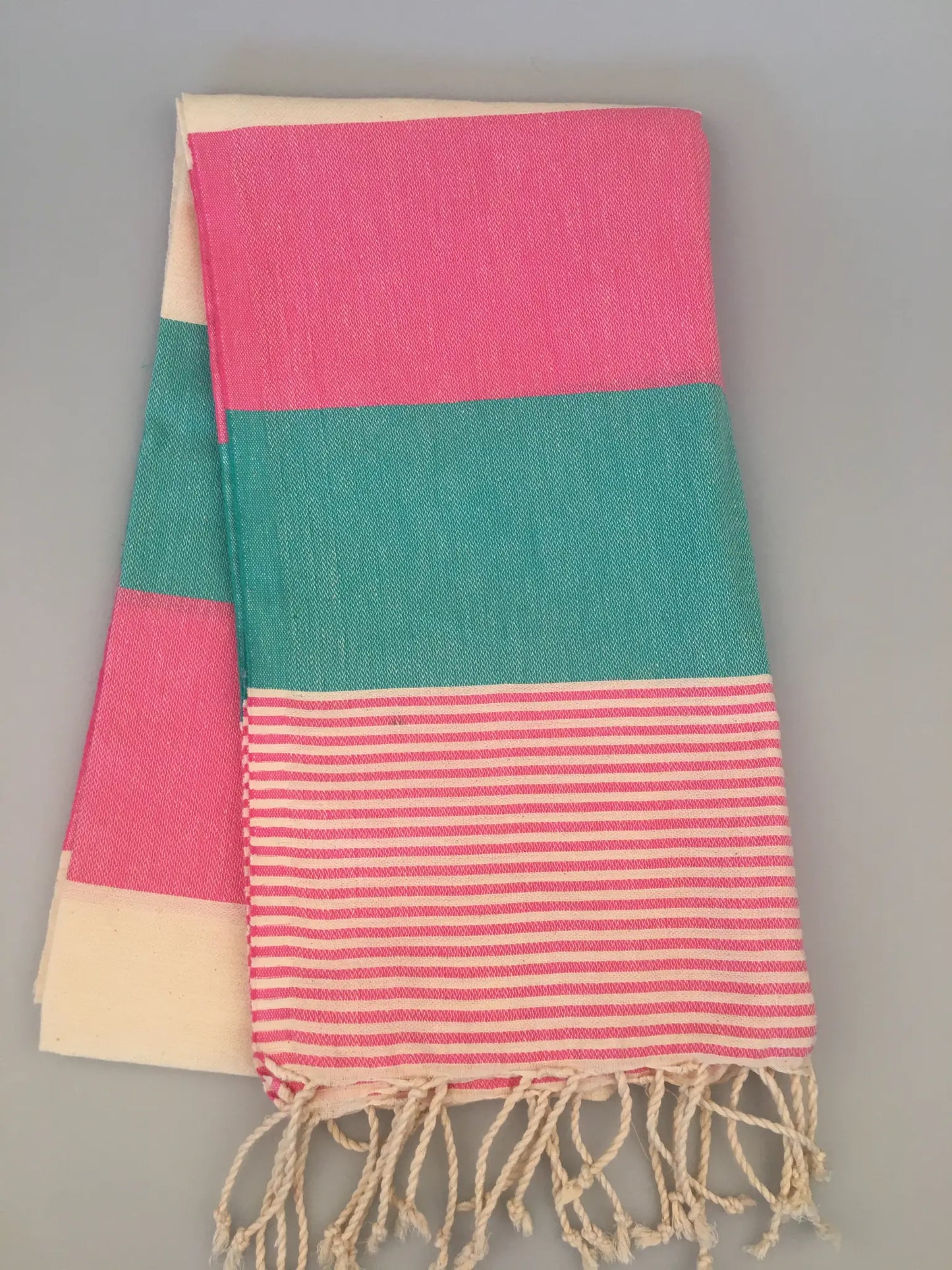 "Turquoise and pink striped Lagina Turkish towel with cream accents and fringed edges, folded on a gray background."