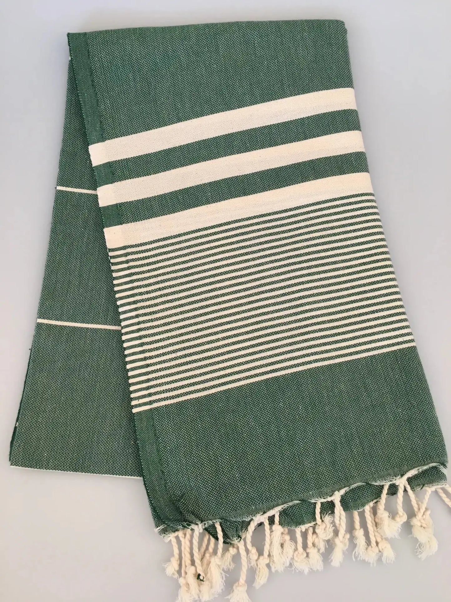 Green Turkish towel peshtemal with white stripes and fringed edges, folded neatly on a light background.