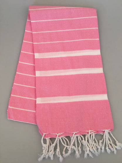"Pink Turkish towel peshtemal with white stripes and fringe edges, laid flat on a gray background."